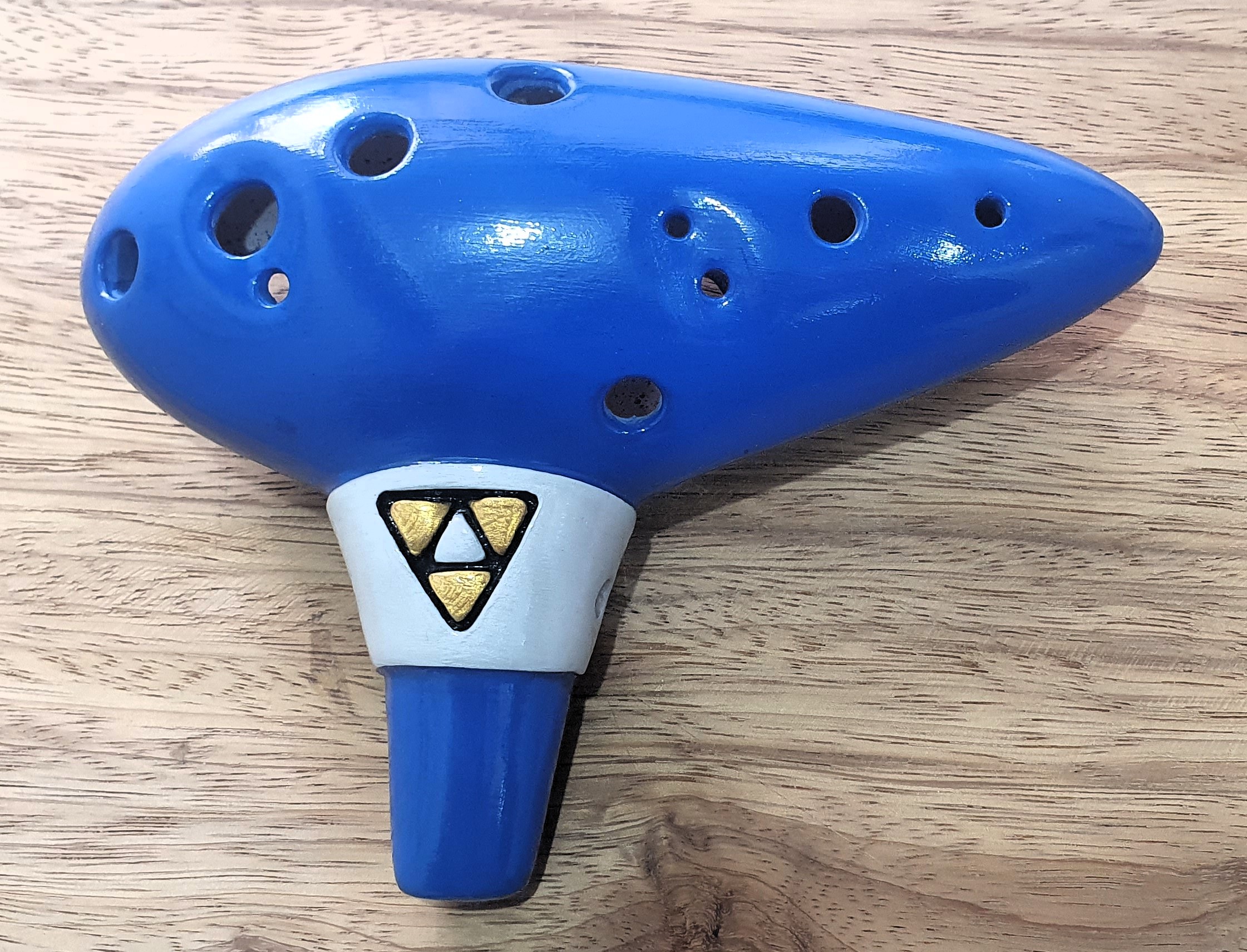 Foto da visão frontal de uma ocarina de 12 furos de cor azul e com símbolo do The Legend of Zelda: Ocarina of Time.
